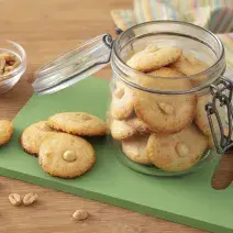 Receita de Crocantinho. Observa-se biscoitos dispostos em uma tábua verde. Ao lado, um pote hermético de vidro com mais biscoitos. Amendoins decoram a foto.