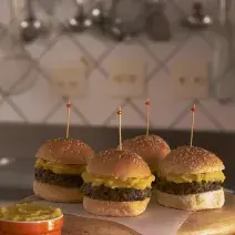 fotografia em tons de cinza e bege de uma bancada marrom vista de frente, contém uma tábua redonda com 4 mini-hambúrgueres com um palito por cima