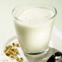 Fotografia em tons de branco e bege em uma bancada de madeira branca, um prato branco, um copo de vidro alto com a bebida com Leite Molico e chá de camomila.