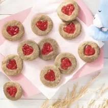 Foto da receita de Biscoitos de Coração. Observa-se uma tábua rosa bebê com os biscoitos dispostos sobe um papel manteiga. Alguns trigos e o ursinho de Mucilon decoram a foto.