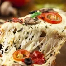 Fotografia de uma pizza com o queijo puxando e bem recheada, como queijo, azeitona, tomates e ricota. A pizza está sobre uma tábua de madeira de toma escuro, e ao fundo, cebola, tomate e champignon.