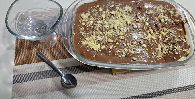 Foto da receita de Pavê de Cupuaçu com Ganache de Chocolate. Observa-se uma travessa retangular com o doce decorado de biscoito picado por cima e ao lado esquerdo tem uma taça individual.