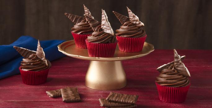 Foto de uma bancada vermelha. Ao centro há um suporte para bolos dourado alto com 3 cupcakes em formas vermelhas e decorados com Nestlé ChocoBiscuits. Na bancada há mais 2 cupcakes, alguns biscoitos e um tecido azul escuro ao fundo.