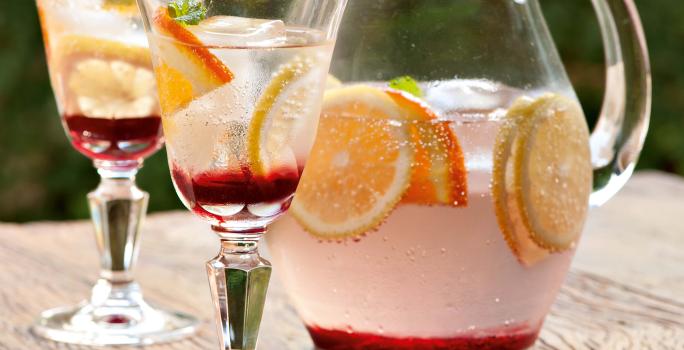 Fotografia em tons de rosa em uma bancada de madeira clara com uma jarra de vidro com o refresco de hibisco e frutas cítricas dentro, decorando com rodelas de laranja. Ao lado, duas taças de vidro com o refresco.