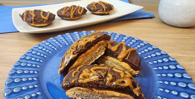 Fotografia em tons de azul em uma bancada de madeira e um prato azul redondo com tortinhas de caramelo salgado com chocolate e aveia em cima dele.