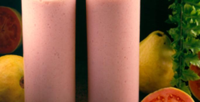 Fotografia em tons de rosa em uma bancada de madeira escura, dois copos de vidro alto com a bebida feita com suco de goiaba e Leite Moça. Ao lado, goiabas e uma goiaba aberta ao meio.