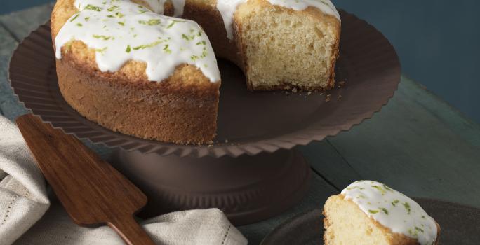 foto em tons de cinza, em uma mesa de madeira contém o bolo de iogurte ao lado um prato marrom com um pedaço de bolo um pano branco e por cima do pano uma espátula.