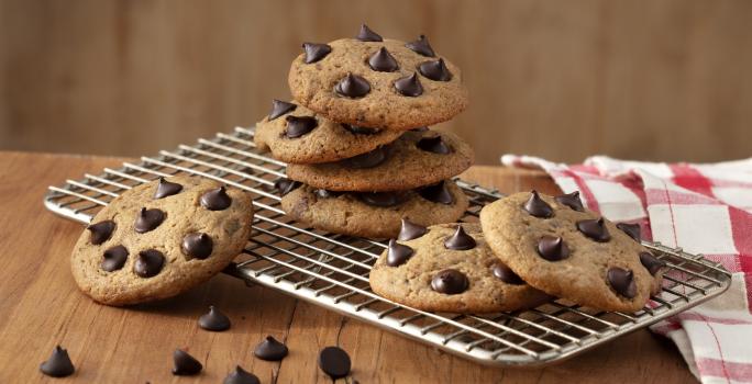 Fotografia em tons de marrom em uma bancada de madeira, um pano xadrez vermelho, um suporte de grelha com alguns cookies com gotas de chocolate em cima dele.