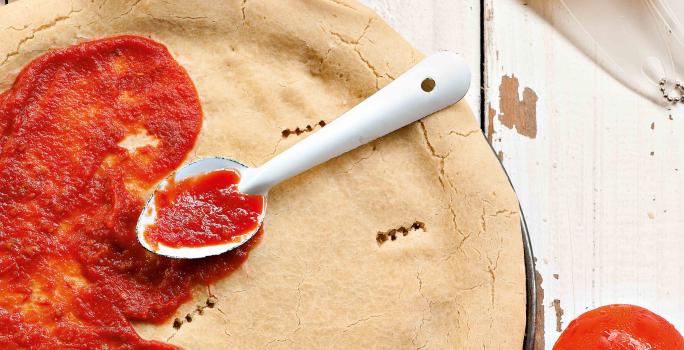 Fotografia em tons de vermelho e branco, de uma bancada de madeira com uma massa redonda de pizza, com molho de tomate pela metade e uma colher prateada. Ao redor potinhos, ovos, tomates e azeite.