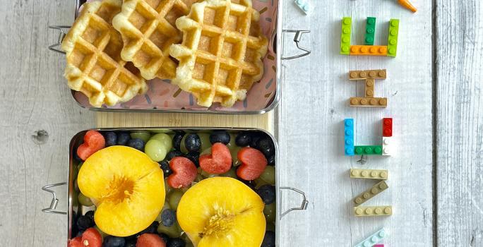 Fotografia em tons coloridos com uma lancheira com waffles e frutas dentro dela. Ao lado letras de brinquedo escrito a palara lanche em uma bancada azul em madeira.