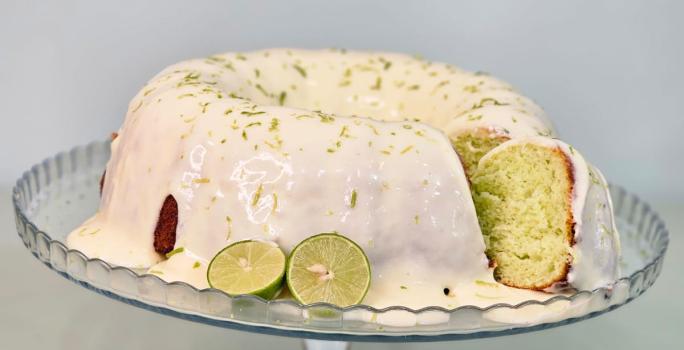 Fotografia em tons de branco com um bolo redondo ao centro. Em cima do bolo existe uma cobertura branca coberta com raspas de limão. Ao lado existe um limão verde cortado ao meio.
