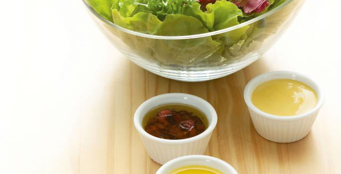 Fotografia em tons de verde em uma mesa de madeira clara com um bowl de vidro com salada de alface e rúcula e ao lado, quatro potinhos brancos com os molhos de morango, mostarda, hortelã e gengibre.