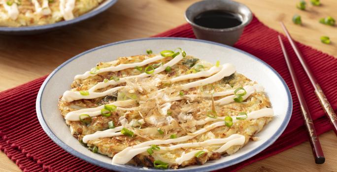 Fotografia em tons de vermelho em uma bancada de madeira com um jogo americano vermelho escuro, um prato branco com detalhes em azul ao centro com o okonomiyaki dentro dele e ao lado, um par de hashi. Ao fundo, um potinho com o molho de soja.