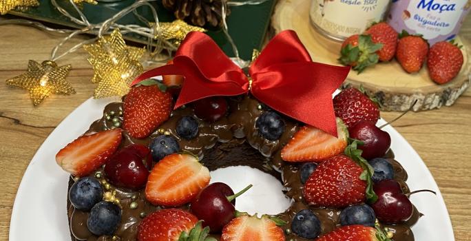 Foto da receita de guirlanda de brownie decorada com frutas vermelhas e decoração de natal servida em um prato grande branco sobre uma mesa de madeira com produtos de leite moça ao fundo