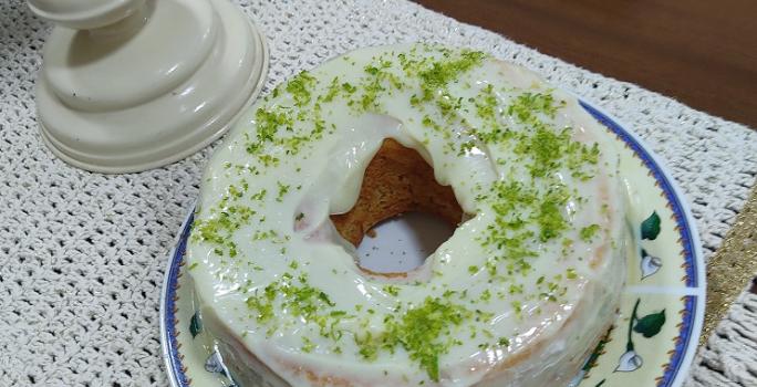 Foto em tons de branco da receita de bolo de nescafé com cobertura de limão servida em uma porção grande sobre um prato decorado em cima de uma mesa de madeira com um pano bordado
