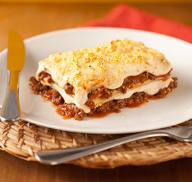 Pedaço de lasanha à bolonhesa sobre um prato branco em uma mesa com garfo e faca apoiados. Pedaço de lasanha à bolonhesa sobre um prato branco em uma mesa com garfo e faca apoiados.