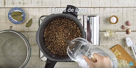 Foto tirada de cima mostra uma panela cheia de feijão cru enquanto alguém despeja água de uma jarra