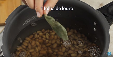 Foto tirada de cima mostra uma panela cheia de feijão cru enquanto alguém despeja água de uma jarra