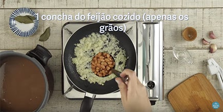 Pessoa despeja uma concha de feijão sobre a frigideira com alho e cebola picados