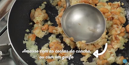 Pessoa usa as costas da concha para amassar feijão na frigideira com alho e cebola
