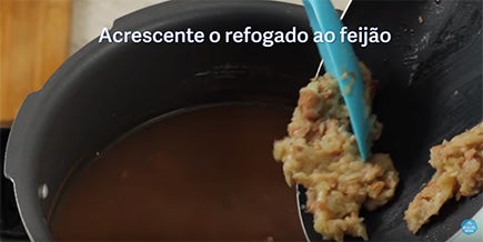 Pessoa despeja feijão amassado com alho e cebola na panela de pressão aberta com feijão cozido