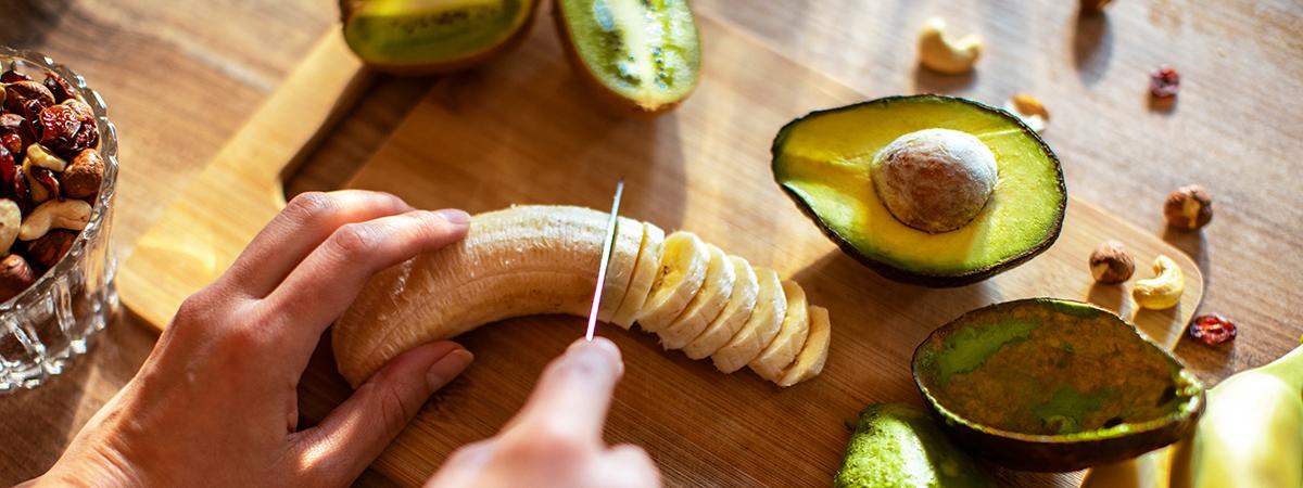 Capa Safra de Março: Descubra 15 receitas equilibradas com as frutas, verduras e legumes do mês! Safra de Março: uma pessoa cortando uma banana, com um abacate cortado do lado