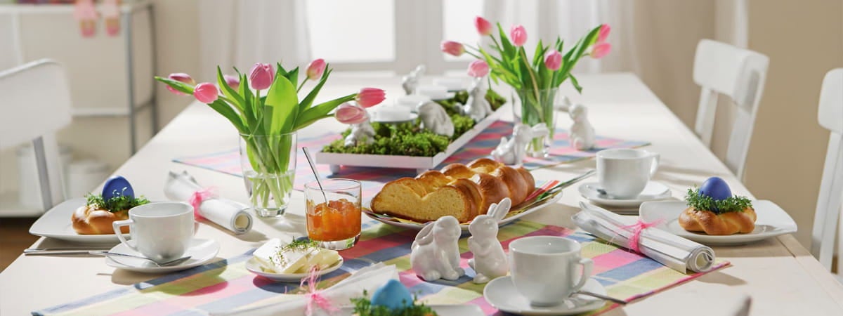 Decoração de Páscoa: Mesa decorada com pratos, talheres, pães, vaso com flores e xícaras