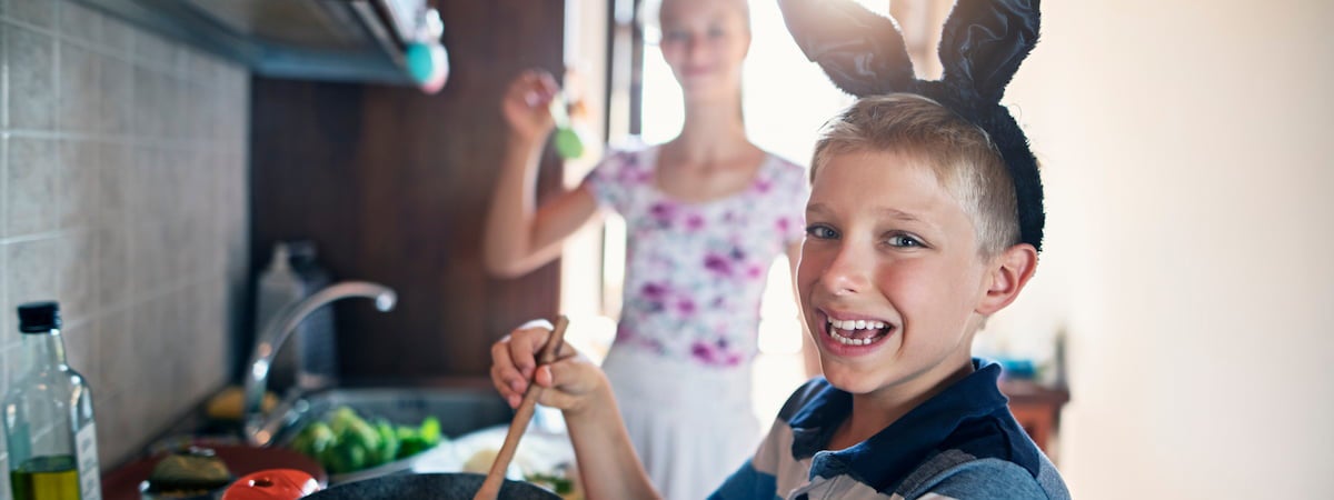 Sexta-Feira Santa: Menino com tiara de coelho sorrindo enquanto segura uma colher ao lado da mãe na cozinha