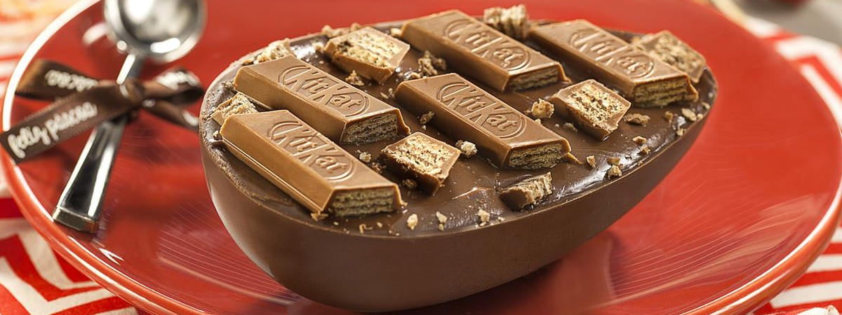 Capa: Ovo de Colher Ovo de colher: Ovo de páscoa com pedaços de chocolate KitKat em cima, em um prato ao lado de uma colher