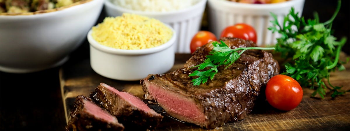 Capa: Acompanhamento para churrasco Carne cortada em três pedaços com tomates em volta, algumas tigelas, em cima de uma mesa de madeira