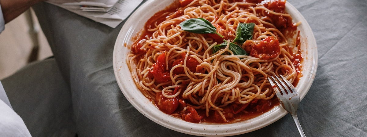 Capa: Almoço rápido Opções de almoço rápido e fácil:Macarrão com molho de tomate no prato com um garfo ao lado em cima de uma mesa com pano cinza