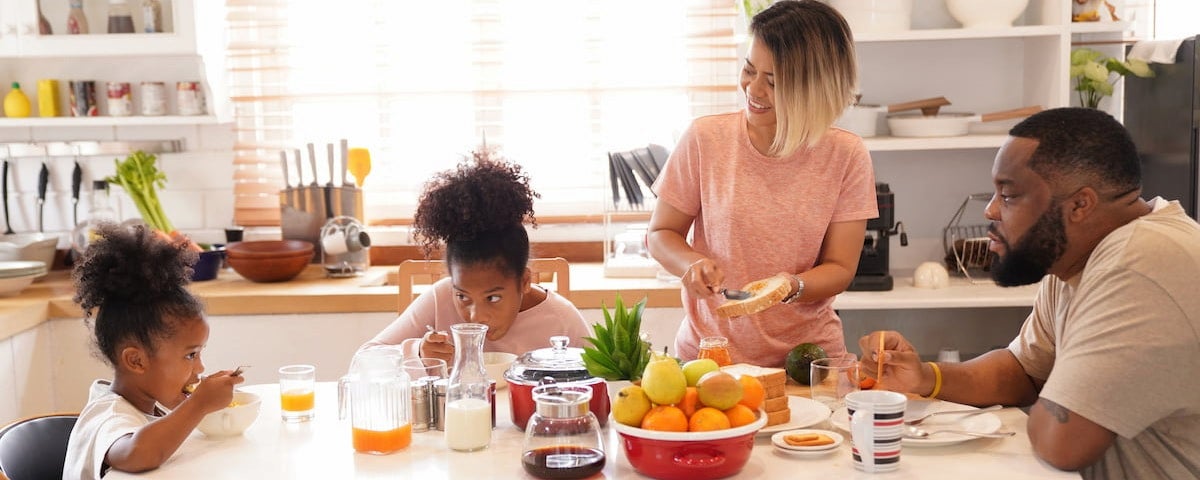 Dicas de café da manhã:Duas crianças negras à esquerda, dois adultos negros à direita, com frutas, leite, pão e café na mesa