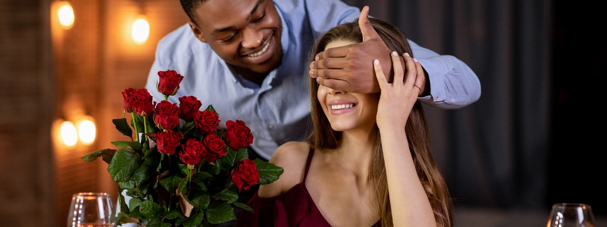 Jantar para o Dia dos Namorados: Homem negro segurando um buquê de flores e com a mão nos olhos de uma mulher branca 