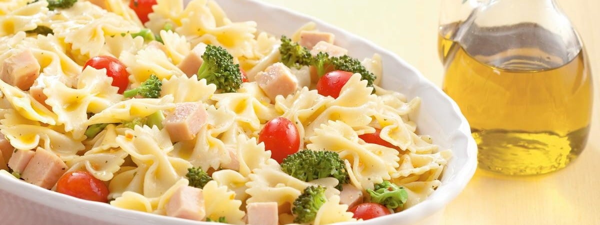 Receitas de saladas: Prato de salada de macarrão com brócolis, peito de peru e tomate cereja, ao lado tem uma garrafa com aze