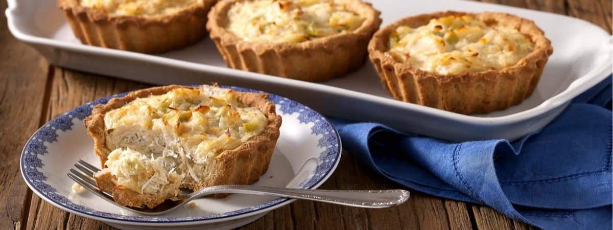 Capa: Alho-poró Quiche de alho-poró sendo cortado sobre um prato com um garfo ao lado. Ao fundo, mais 3 quiches inteiros