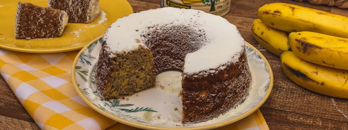 Bolo de banana ôrganico Ninho Bolo de banana com um pedaço cortado, à direita 5 bananas e, à esquerda os pedaço do bolo que foram cortados sob um prato