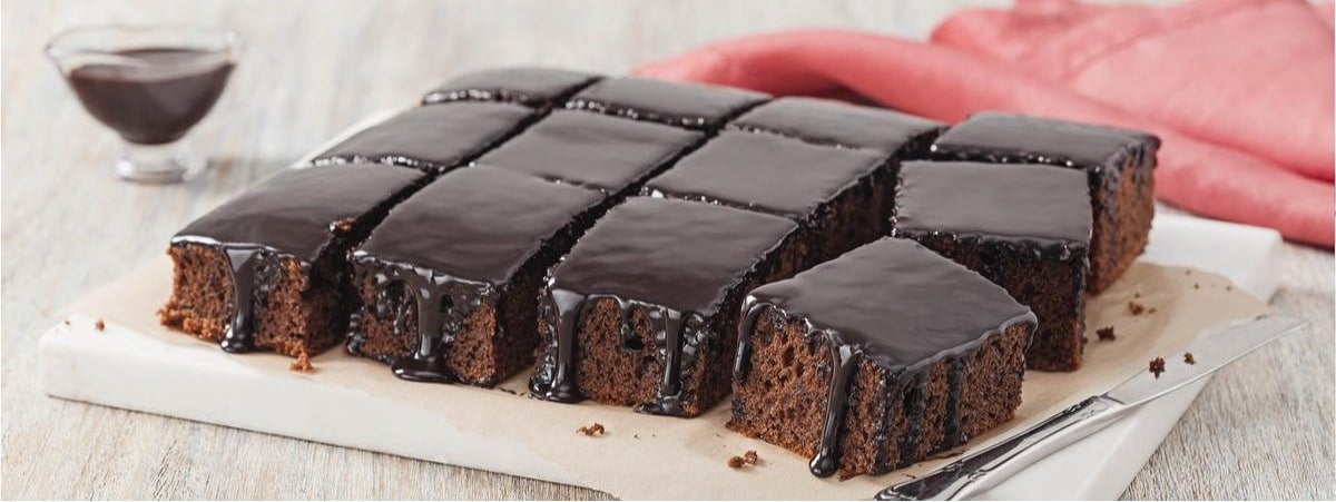 Bolo de chocolate Prato com um bolo de chocolate cortado em pedaços quadrados e uma faca, ao fundo um pano vermelho e um potinho com chocolate