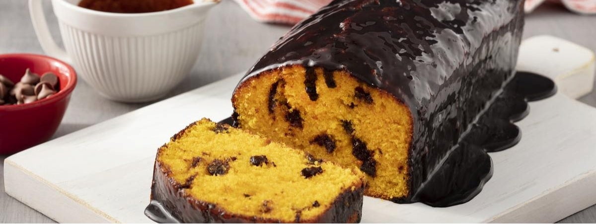Capa: Bolo fofinho Bolo de cenoura com cobertura de chocolate em cima de um prato, à esquerda pote com gotas de chocolate