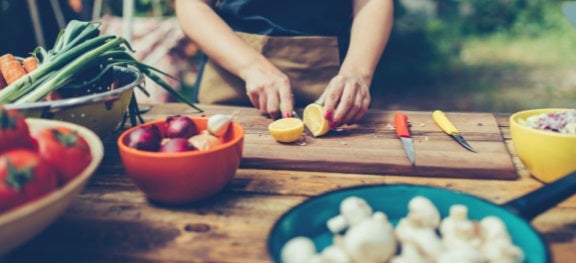 Pessoa cortando uma laranja ao meio. Perto dela, bowls com cebolas, tomates, cogumelos e cenouras. Pessoa cortando uma laranja ao meio. Perto dela, bowls com cebolas, tomates, cogumelos e cenouras.