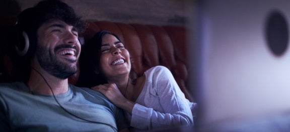 Casal assistindo TV e sorrindo. O rapaz está com fones de ouvido e a moça está apoiada nele. Casal assistindo TV e sorrindo. O rapaz está com fones de ouvido e a moça está apoiada nele.