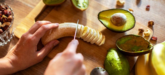 Na imagem, vemos uma mesa de madeira, sobre ela temos uma tábua de corte também em madeira. Sobre ambas, vemos uma pessoa cortando frutas como banana, abacate e no canto superior esquerdo é possível ver uma vasilha de vidro com castanhas. Na imagem, vemos uma mesa de madeira, sobre ela temos uma tábua de corte também em madeira. Sobre ambas, vemos uma pessoa cortando frutas como banana, abacate e no canto superior esquerdo é possível ver uma vasilha de vidro com castanhas.