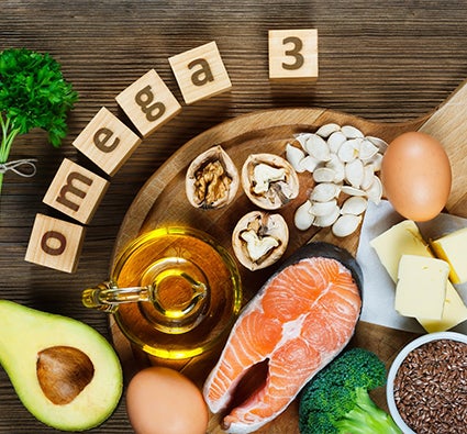 Alimentos ricos em ômega-3 sobre uma tábua: salmão, abacate, ovos, oleaginosas, azeite, manteiga e brócolis.