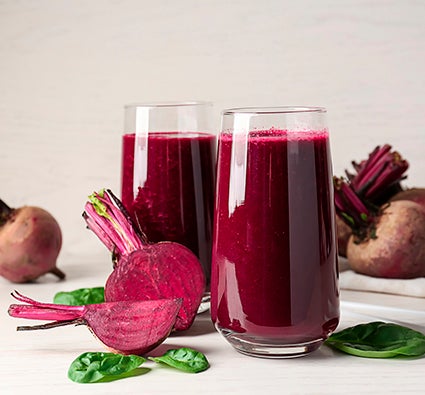 Copos com suco de beterraba, rodeados por beterrabas frescas cortadas e folhas verdes sobre superfície clara.