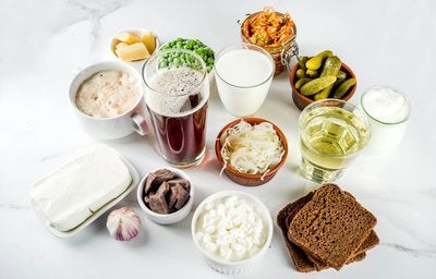 Alimentos ricos em probióticos e prebióticos sobre mesa branca: picles, kefir, cottage, iogurte, kombucha e pães integrais.
