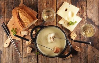 Imagem de uma mesa de madeira com uma panela de fondue e acompanhamentos como pães, queijos e vinho.