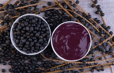 Tigela com polpa de açaí e tigela com frutas de açaí inteiras, sobre ramos com frutos dispostos em superfície clara.
