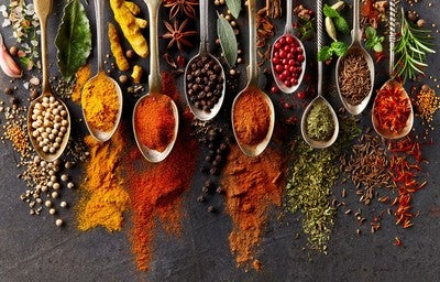 Colheres com diferentes temperos e especiarias coloridas espalhadas sobre superfície escura.