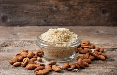 Pote de vidro com farinha de amêndoas, cercado por amêndoas cruas sobre superfície de madeira rústica.