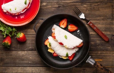 Duas tapiocas recheadas com frutas em uma frigideira sobre uma mesa de madeira. Ao lado, um prato com outra tapioca.