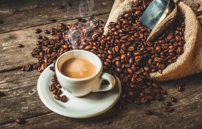 Xícara de café quente sobre mesa rústica com grãos de café espalhados e saco de juta ao fundo.